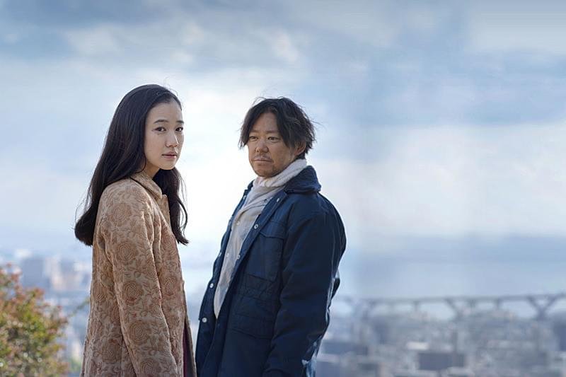 高台の街並みを背に並んで立つ十和子と陣治(映画『彼女がその名を知らない鳥たち』)
