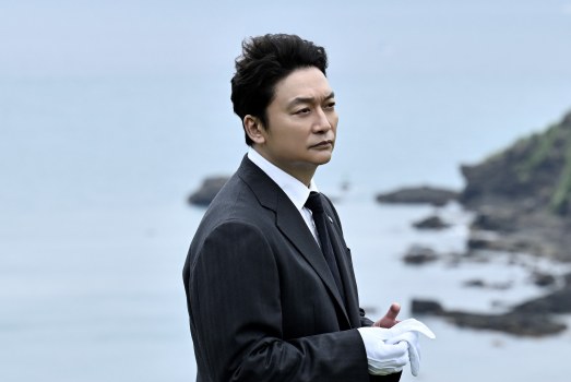海辺に立つ黒いスーツに白い手袋の男性(連続ドラマW「虚ろな十字架」第1話の場面)